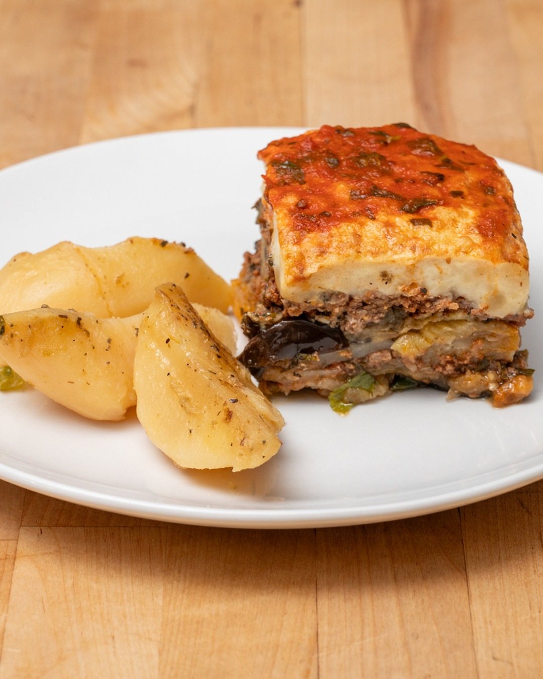 A square of moussaka with layers of eggplant, seasoned meat and béchamel, next to lemon potatoes on a white plate